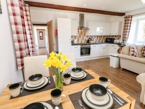 A dining table set with plates, bowls, and cups, a vase with yellow flowers, and a kitchen in the background. - Stable Cottage, Gatcombe (Newport)