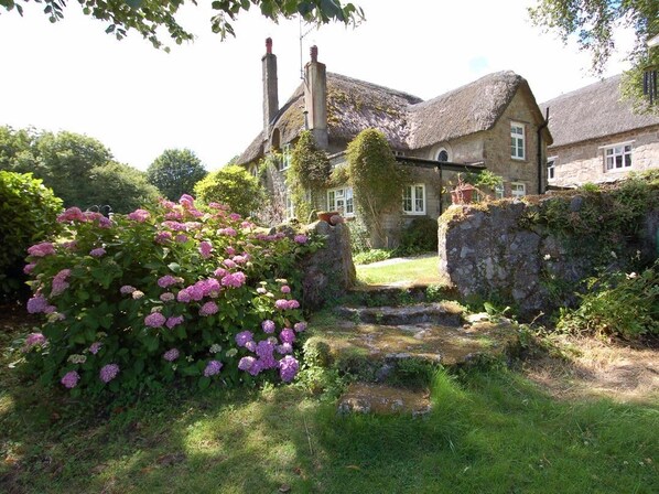 A stone house with a thatched roof, surrounded by lush greenery and flowering plants.