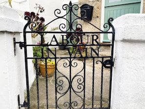 A wrought iron gate with 'LABOUR VAN' engraved on it, leading to a white wall with a green door, potted plants, and a mailbox. - Labour in Vain, St Ives (St. Ives)