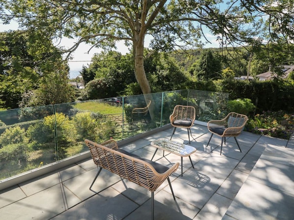 Un patio al aire libre con mobiliario de mimbre, una mesa de vidrio y vistas a un jardín exuberante y colinas lejanas.