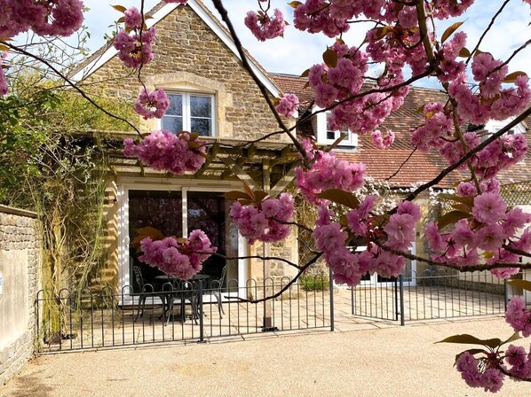A stone building with a covered entrance, a fenced patio, and pink flowering branches in the foreground.