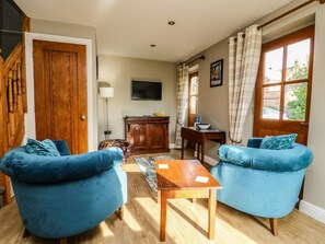 A living room with two blue sofas, a wooden coffee table, a TV mounted on the wall, and a window with curtains. - Decca Cottage Nidd Grange, York (York)