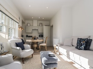 Ein modernes Wohnzimmer mit Sofa, Sessel, Hocker und einem Essbereich mit Tisch und Stühlen.