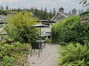 Ein Garten mit Tisch und Stühlen, umgeben von dichtem Grünwerk und einem Gebäude im Hintergrund.