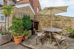 Eine Steinsäule, ein Holztisch, Stühle und Kübelpflanzen in einem Innenhof.