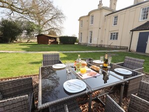 An outdoor dining area with wicker chairs and a glass table set for a meal, surrounded by a well-maintained lawn and a large building in the background. - Moortown House, Caistor (Market Rasen)
