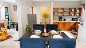 A dining area with a table set for four, a kitchen with wooden cabinets, and a living room with a sofa and a plant. - Peaceful 2-bedroom house in charming Arusha for your getaway (Arusha)