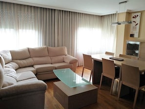 Een woonkamer met een bank, een glazen salontafel, een eettafel met stoelen en een keuken met kasten.