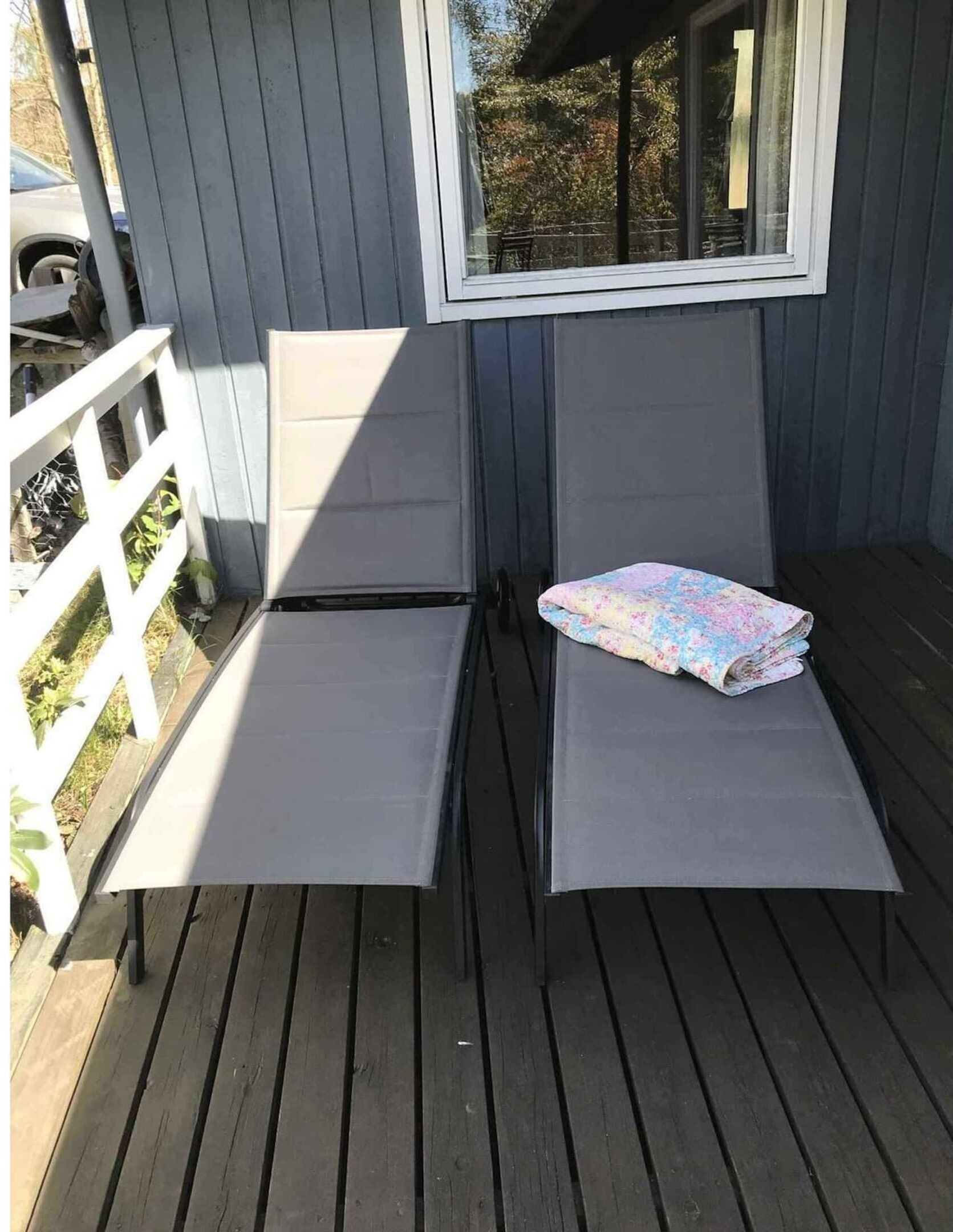 A small outdoor deck with two chairs and a table, a window, and a view of trees.