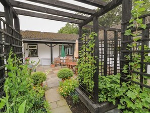 Un jardin agrémenté d’une pergola en bois, une terrasse avec une table et des chaises, et un bâtiment blanc avec une porte verte.