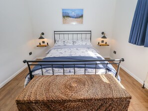 Una cama con estructura metálica, una mesita de noche con lámpara, un estante en la pared y un banco de mimbre.