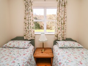 A room with two beds, a nightstand with a lamp, and a window with curtains.
