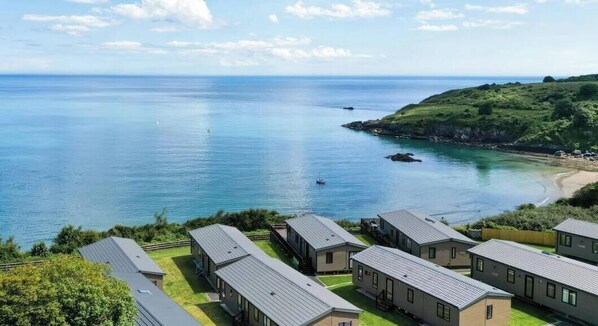 Vista costera con una hilera de cabañas modernas de una planta y techos planos, ubicadas en una zona verde con vistas despejadas al mar y un paisaje montañoso al fondo.