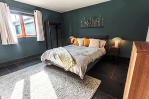 Un dormitorio con cama, lámparas de noche, una ventana con cortinas, una obra de arte en la pared y suelo oscuro.
