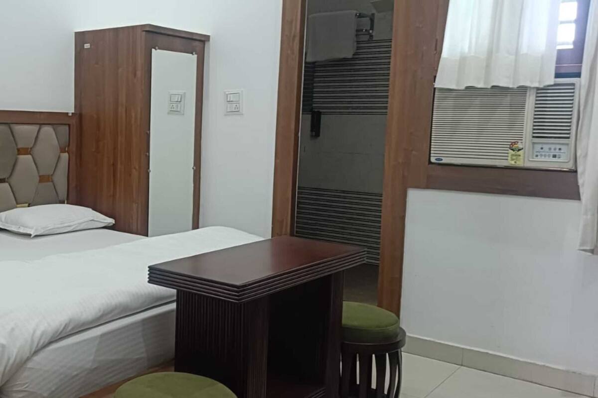 Un dormitorio con una cama, una mesita de noche de madera, un espejo, un taburete verde y una ventana con cortinas.