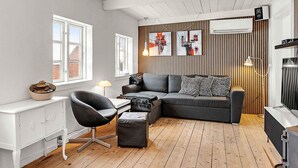 Ein modernes Wohnzimmer mit einer Couch, einem Sessel und einem kleinen Tisch. Es gibt zwei Fenster, einen Deckenventilator und Kunstwerke an der Wand.