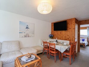 A cozy living area with a sofa, dining table, and chairs, a television mounted on a wooden wall, and a painting of a lighthouse.