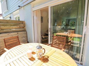 An outdoor wooden table with chairs, a glass door leading to an indoor area, and a view of a garden through the door.