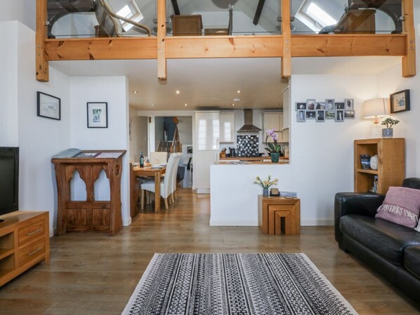 A spacious living area with a high ceiling, wooden beams, a black leather sofa, a patterned rug, and a kitchen in the background.