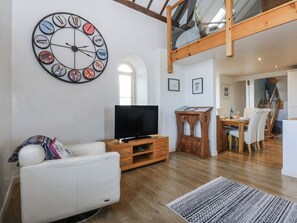 A living room with a large wall clock, a flat-screen TV, a white sofa, a wooden coffee table, and a dining area with a wooden table and chairs.