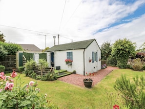 Une maison blanche avec un toit vert, un petit jardin devant avec un chemin de gravier et un autre jardin avec diverses plantes et fleurs.