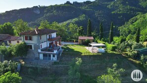 Una casa de dos pisos con balcón, rodeada de abundante vegetación y colinas.