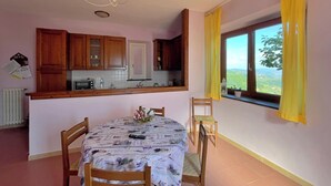 Un comedor con una mesa dispuesta para dos, armarios de madera arriba, una ventana con cortinas amarillas y vistas al exterior con follaje.