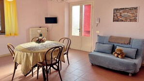 Un comedor con una mesa y sillas, un sofá con un oso de peluche, una araña, un televisor y una puerta con paneles de vidrio esmerilado.