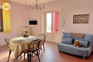 A dining area with a table and chairs, a sofa with a teddy bear, a chandelier, a TV, and a door with frosted glass panels. - Tuscan Olive Grove Retreat with Hot Tub & Medieval Village Views (Toscana)