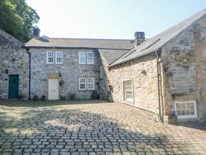 A stone building with a cobblestone courtyard, a green door, and a window with a flower box. - Querc, Stoney Middleton (Hope Valley)