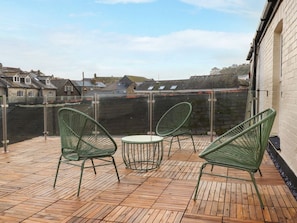 Une terrasse en bois avec des chaises vertes et une petite table, donnant sur un mur de pierre et des bâtiments.