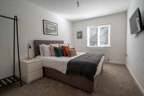 A bedroom with a bed, bedside tables, a radiator, a window, and a TV mounted on the wall. - The Cove, Kessingland (Lowestoft)