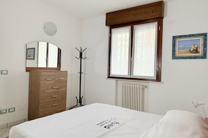 A bedroom with a bed, a wooden dresser, a coat rack, a window with curtains, a radiator, and a framed picture on the wall.