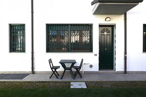 A white building with a black door, two windows with metal bars, a table with chairs, and a small outdoor area with a mat.