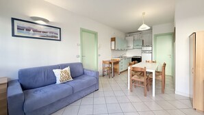 A living room with a blue sofa, a dining table with chairs, a kitchen area, and light green doors.