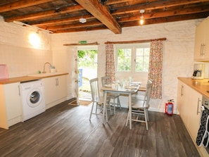 A kitchen with a wooden ceiling, a washing machine, a dining table with chairs, and a window with floral curtains.