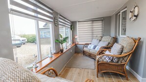 Een zonnige kamer met een rieten bank, een fauteuil met kussens, en een raam met uitzicht op een auto en groen buiten.