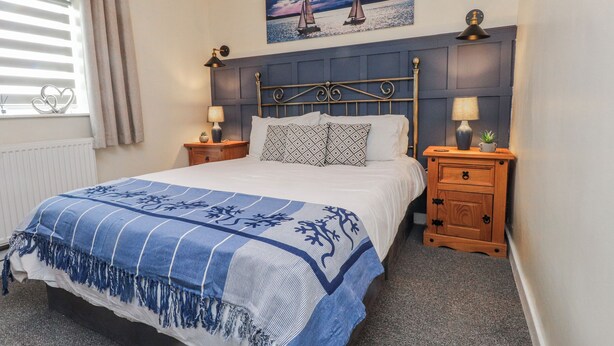 Een slaapkamer met een bed, twee nachtkastjes, een hoofdbord, een raam met jaloezieën, een schilderij van zeilboten en een radiator.