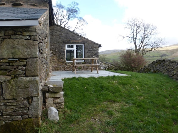 A stone building with a wooden bench outside, a stone wall, and a grassy area.