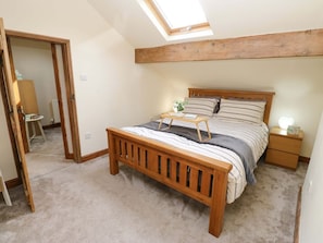 Une chambre avec un lit en bois, une table de chevet, une commode, un puits de lumière et une porte donnant sur une autre pièce.