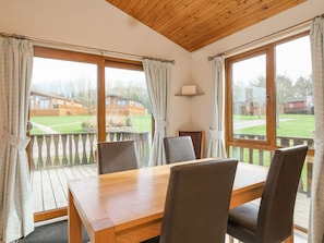 A room with a wooden table and chairs, large windows with curtains, and a view of a wooden deck and buildings.