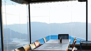 A long dining table with chairs, large windows, and a view of mountains. - Shelar Agro Tourism and Holiday Homes (Mahabaleshwar)