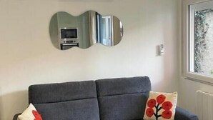 En grå sofa med to dekorative puder, et rundt sofabord med en plante, og et spejl på væggen over en hvid aircondition.