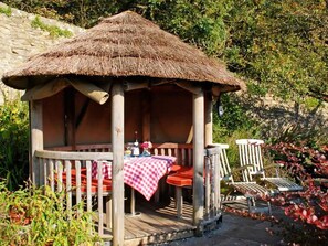 A wooden gazebo with a thatched roof, a table with a checkered tablecloth, and red chairs.