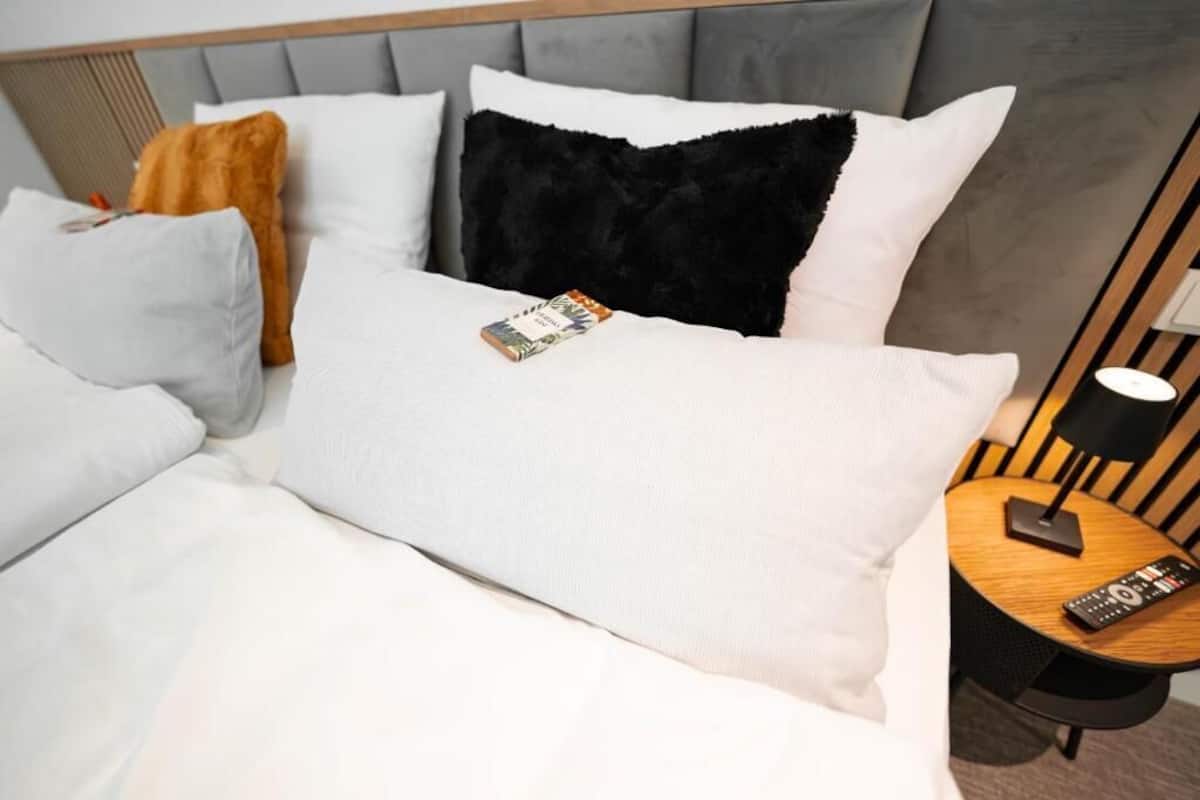 Una habitación de hotel con una cama, almohadas, un control remoto y una lámpara sobre una mesita de noche.