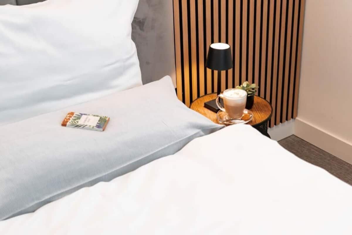 Una cama bien hecha con sábanas blancas, una mesita de noche con una lámpara, una taza de café y un libro.
