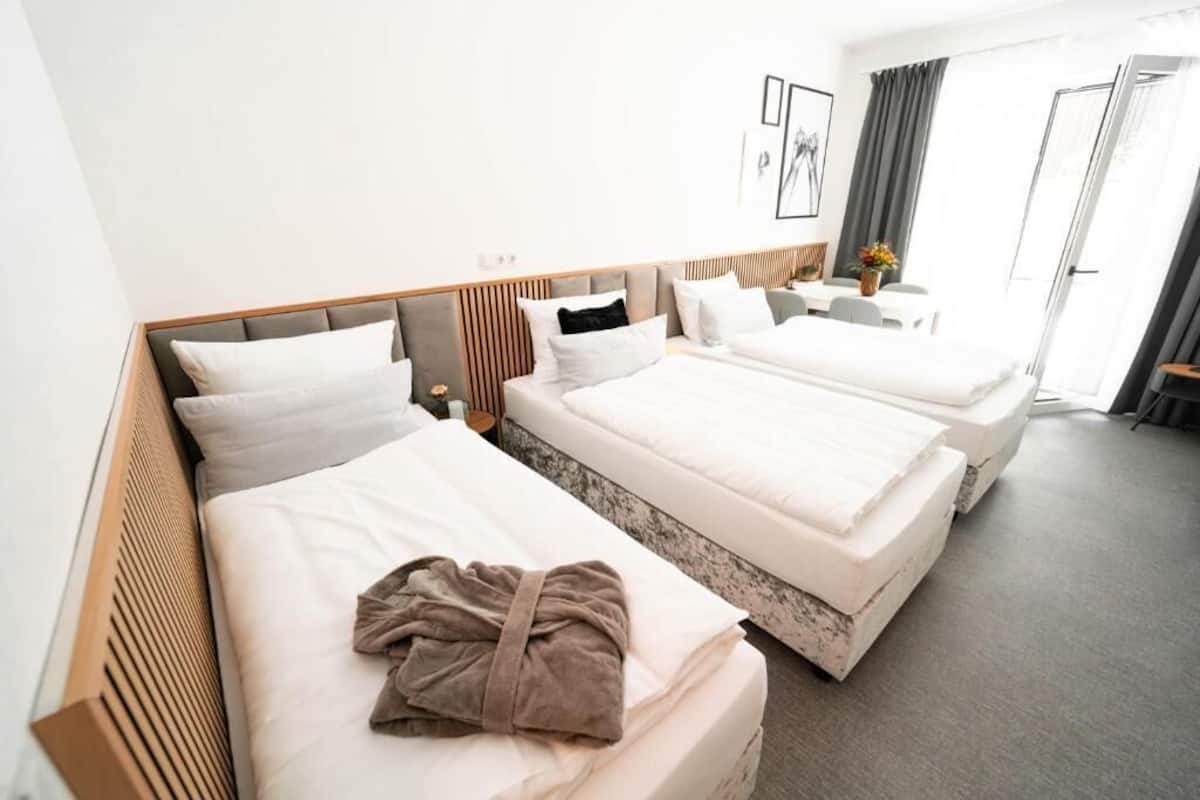 Habitación de hotel con tres camas, una mesita con un florero y vistas al exterior.