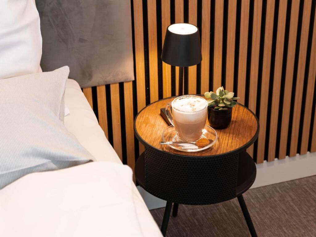 A bed with white bedding, a round wooden side table with a black lamp, a cup of coffee, and a small potted plant.