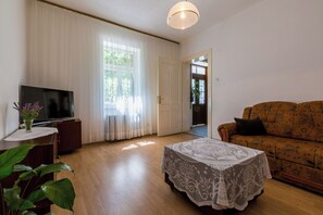 A living room with a TV, a table with a lace tablecloth, a sofa, and a door leading to another room. - Vacation home in Jadranovo with Mediterranean garden & balcony (Jadranovo)