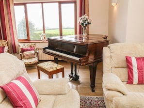 Un salon agrémenté d’un magnifique piano à queue, de fauteuils à motifs floraux et offrant une vue sur l’extérieur grâce à de grandes fenêtres.